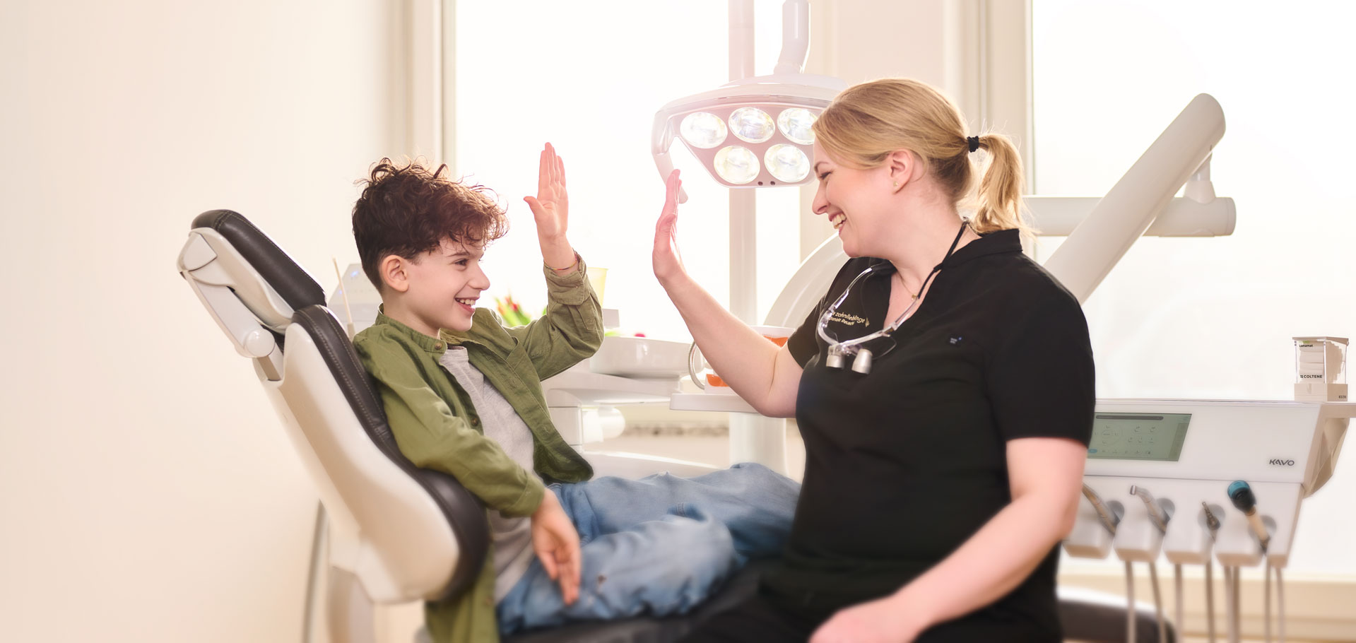Zahnärztin Dr. Daniela Reuschl gibt einem Jungen in grünem Hemd ein High Five, während dieser auf dem Behandlungsstuhl ihrer Zahnarztpraxis sitzt