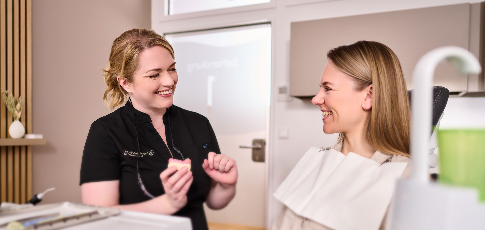 Im Behandlungsraum der Zahnarztpraxis Zahnlieblinge erklärt Zahnärztin Dr. Reuschl einer Patientin etwas mithilfe des Modells eines Gebisses