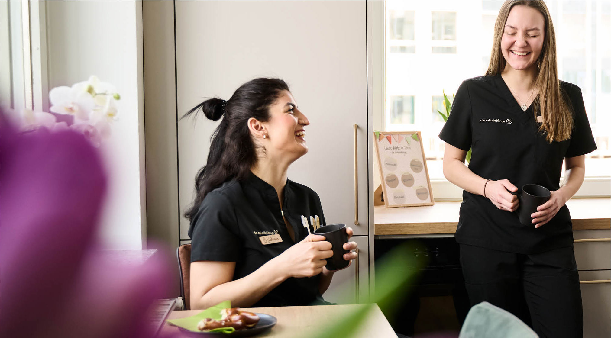 Zwei Mitarbeiterinnen der Zahnarztpraxis lachen gemeinsam im Aufenthaltsraum, während sie einen Kaffee trinken